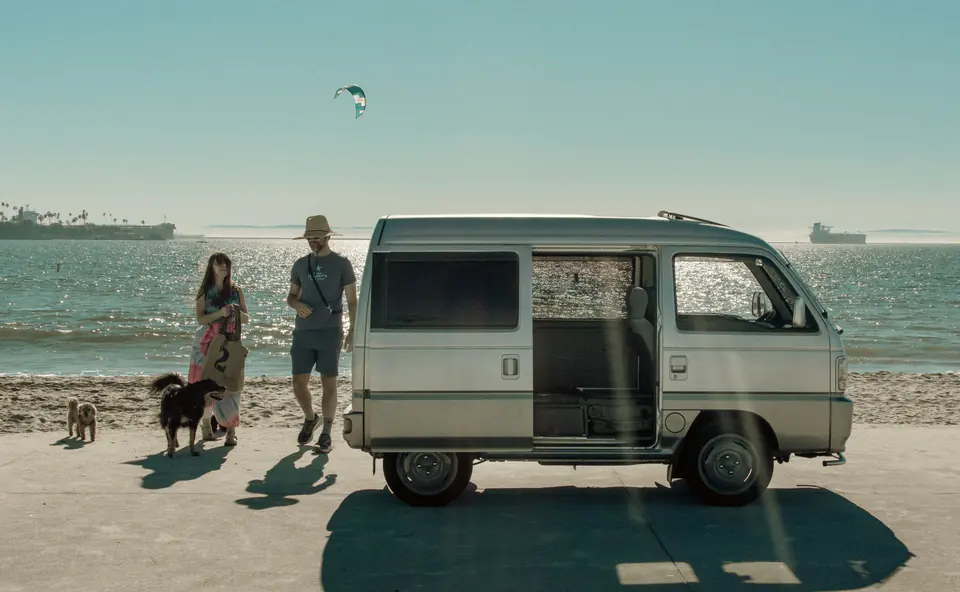 Couple walking their dogs past a silver Honda Acty Street Kei Van parked on the beach
