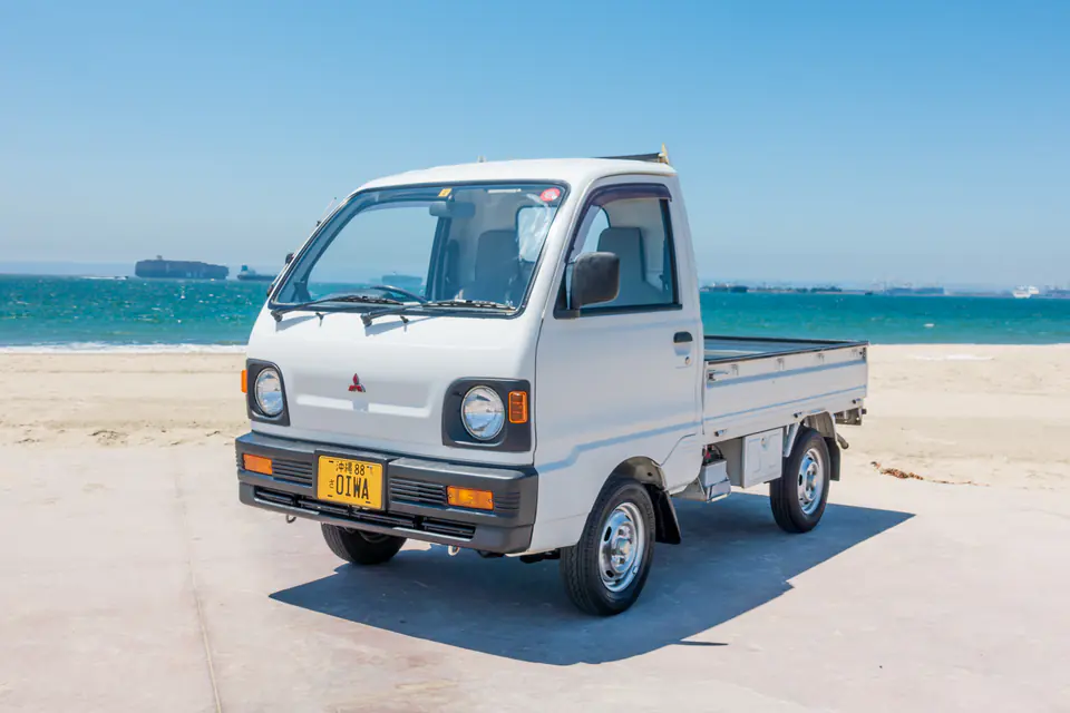 White 1990s Mitsubishi Minicab Kei Truck with round headlights parked on the beach