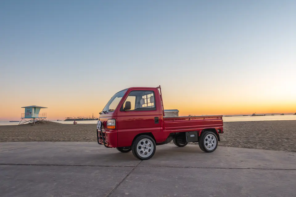 Side profile of a red Honda Acty mini truck on the beach at sunset with a lifeguard tower behind it
